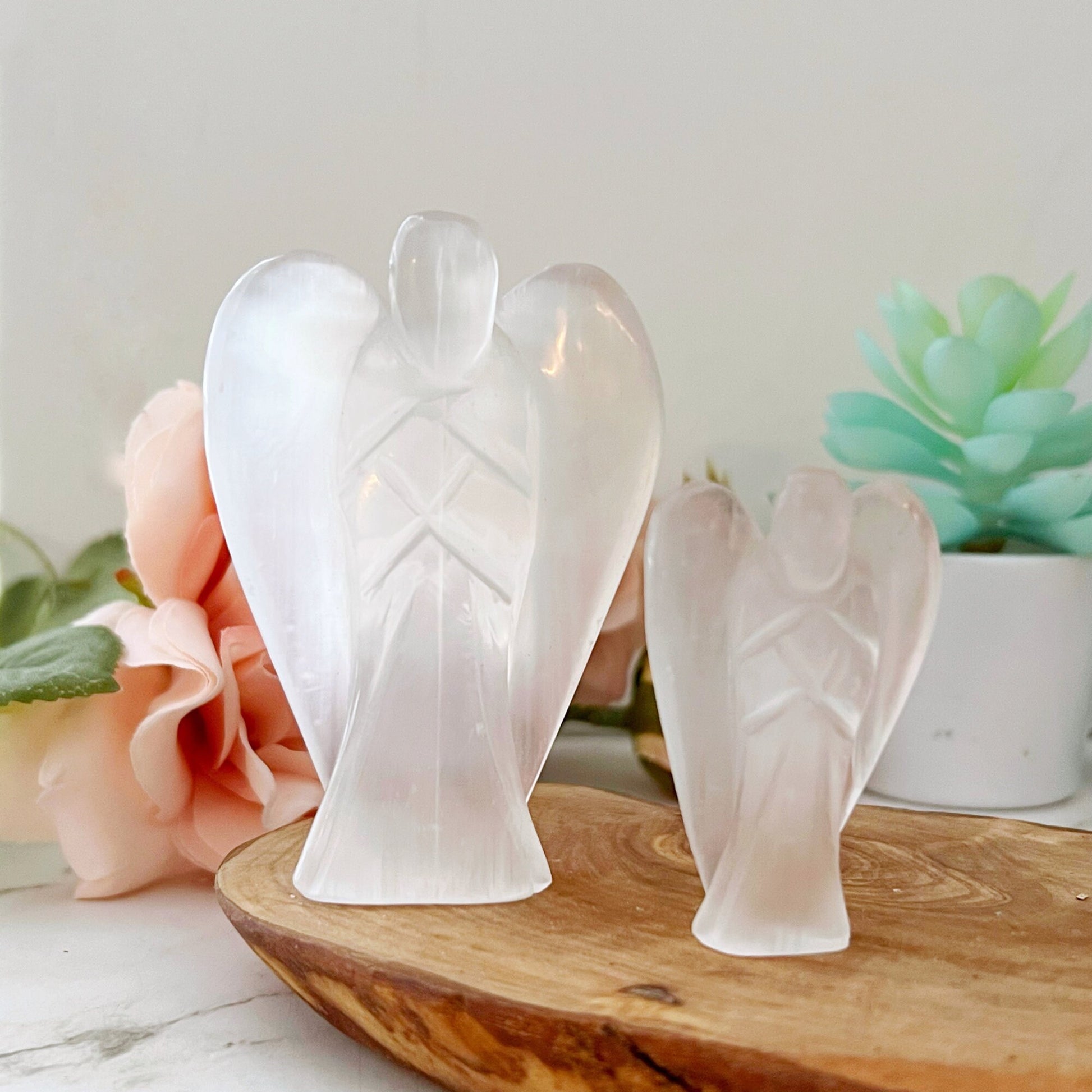 Three 1.5" or 3" Selenite Crystal Angels of varying sizes are displayed on a wooden tray. The background includes soft-focus flowers and greenery, creating a serene and delicate atmosphere. The figurines have smooth, translucent surfaces that reflect light beautifully, symbolizing spiritual enlightenment.