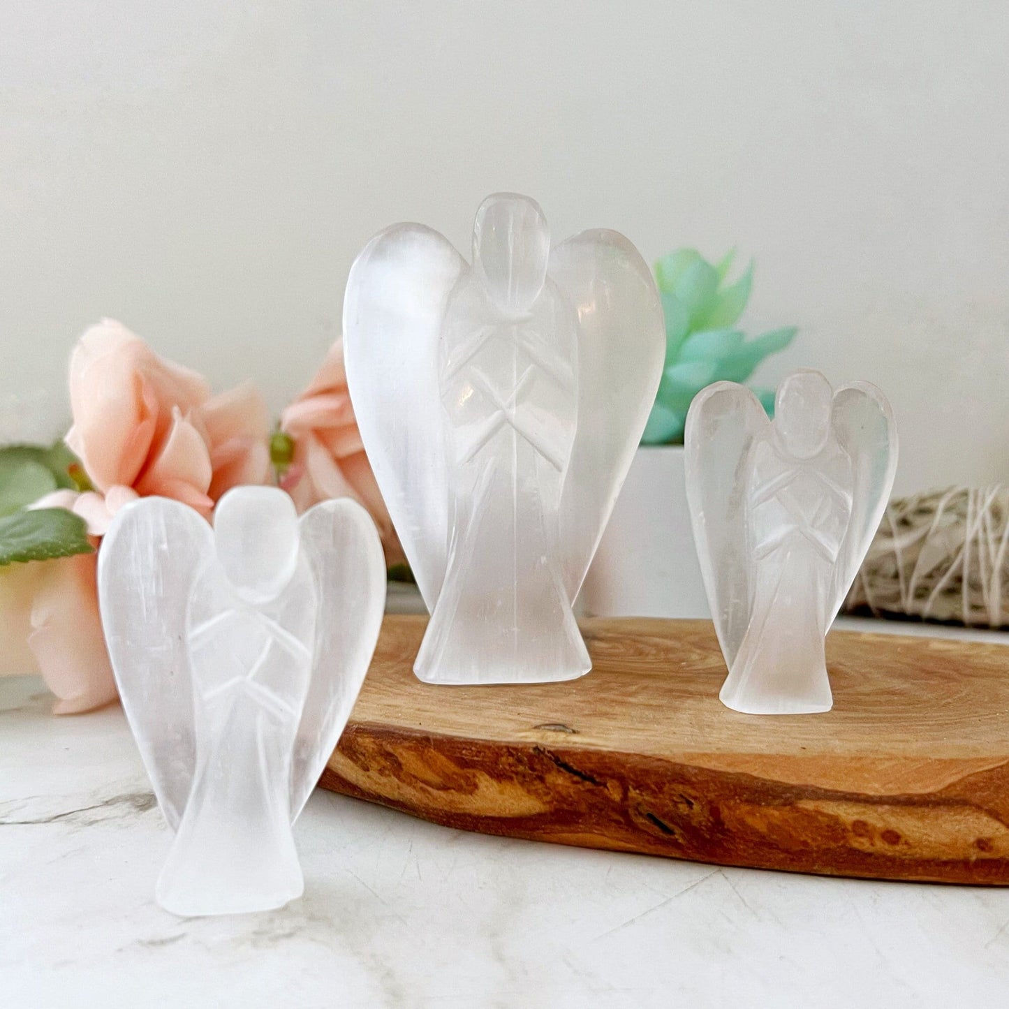 Three 1.5" or 3" Selenite Crystal Angels of varying sizes are displayed on a wooden tray. The background includes soft-focus flowers and greenery, creating a serene and delicate atmosphere. The figurines have smooth, translucent surfaces that reflect light beautifully, symbolizing spiritual enlightenment.