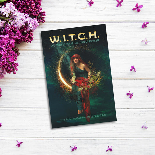 A book titled "WITCH Woman in Total Control of Herself Oracle” lies on a white wooden surface surrounded by purple flowers. The cover art, reminiscent of the W.I.T.C.H. Card Deck, features a mystical woman in a red headscarf holding a crescent moon against a backdrop of greenery and glowing light.