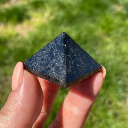 A person holds a small, 1" Mini Black Tourmaline Pyramid against a blurred background of green grass. The smooth, black stone with slight white veining is visible in daylight. Known for its protective properties, it transmits calming energy through the thumb and part of the index finger that are also visible.