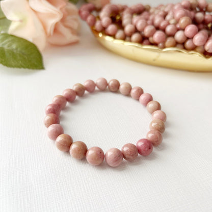 An 8mm Rhodonite Beaded Bracelet is displayed in the foreground on a white surface. Behind it, there is a decorative dish filled with similar pink beads, and a soft-focus bouquet of light pink roses with green leaves in the background, highlighting the metaphysical properties of Rhodonite gemstones.