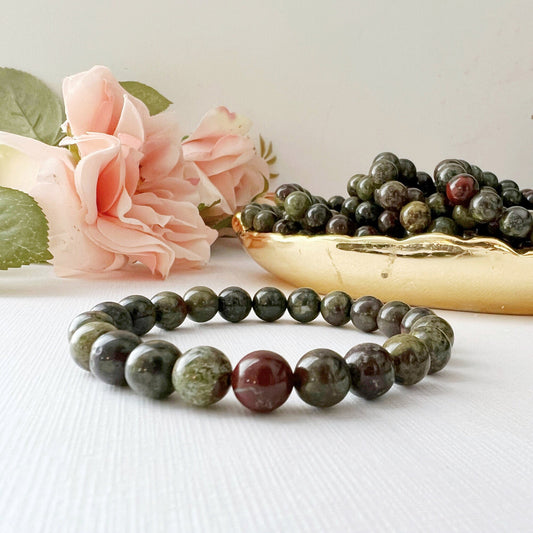 An 8mm Dragons Blood Jasper Beaded Bracelet made of dark green and red stones is displayed on a white surface. In the background, there are pink roses and a golden bowl filled with similar beads. The scene exudes natural beauty, creating a serene and elegant atmosphere.