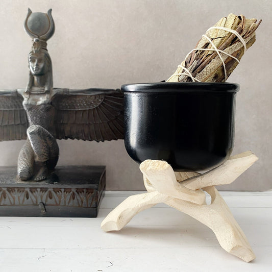 An altar with an Egyptian-style statue of a winged deity stands beside a Black Soapstone Bowl on a wooden stand. The cauldron contains a bundle of sage wrapped in string, and burning incense adds to the mystical atmosphere. The scene is set against a neutral background.