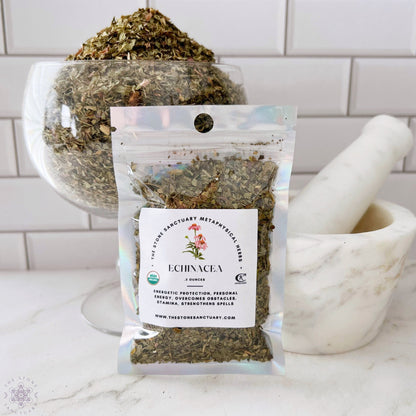 A small clear bag labeled "Echinacea Herb" sits on a white surface in front of a bowl filled with dried leaves. A white mortar and pestle are placed to the right. The background features white tiles, emphasizing the natural, organic non-GMO essence of this metaphysical herb.