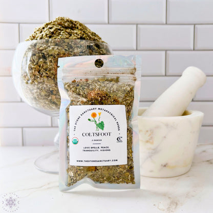 A packet of Coltsfoot Leaf is displayed in front of a bowl filled with more of the same herbs and a white marble mortar and pestle. The packet label mentions metaphysical uses like love spells, peace, tranquility, and visions. The setup is on a white countertop.