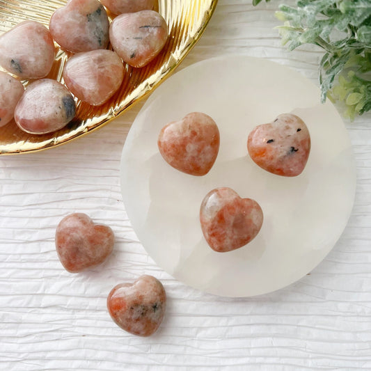 Heart-shaped, peach-colored crystals are displayed on a white textured surface. Four 1" Mini Sunstone Heart crystals are arranged on a round white plate, with a golden dish holding several more 1" Mini Sunstone Hearts in the background. Green foliage partially visible at the top right adds a natural touch.