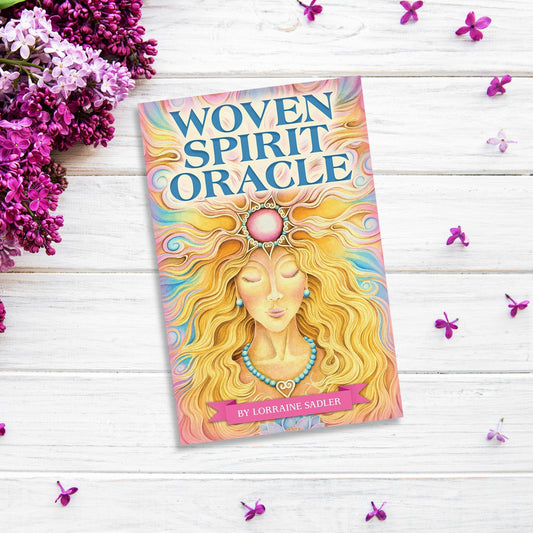 A colorful book titled "Woven Spirit Oracle" by Lorraine Sadler lies on a white wooden surface surrounded by vibrant purple flowers. The cover features ethereal imagery of a serene woman with flowing hair adorned in spiritual symbols, reflecting the essence of this oracle card deck titled *Woven Spirit Oracle*.