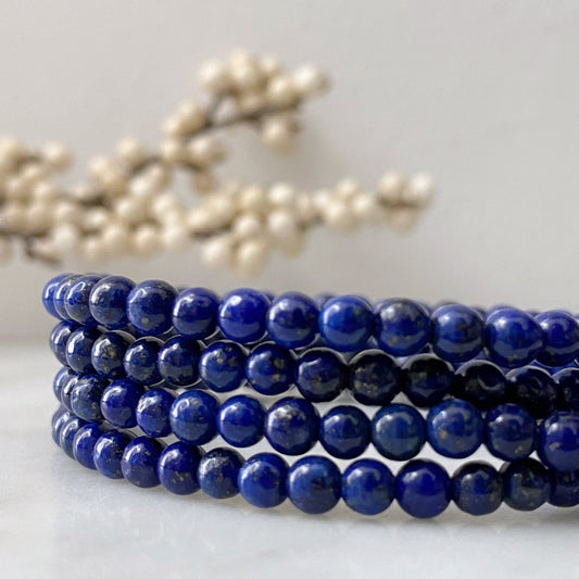 A close-up shot of a 4mm Lapis Lazuli Beaded Bracelet composed of multiple strands of polished, deep blue beads. In the blurred background, delicate white berries are visible, adding a soft, natural accent to the image. The bracelet is placed on a light, neutral surface, radiating Reiki energy for spiritual awareness.