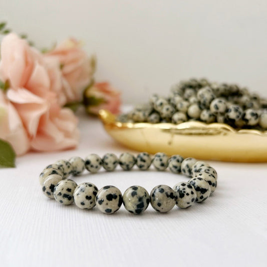 An 8mm Dalmatian Jasper Beaded Bracelet made of beaded gemstone beads is placed on a white surface. In the background, there's a bowl filled with similar beads and some soft pink flowers, adding a touch of elegance to the composition. This piece exemplifies the beauty of metaphysical jewelry.