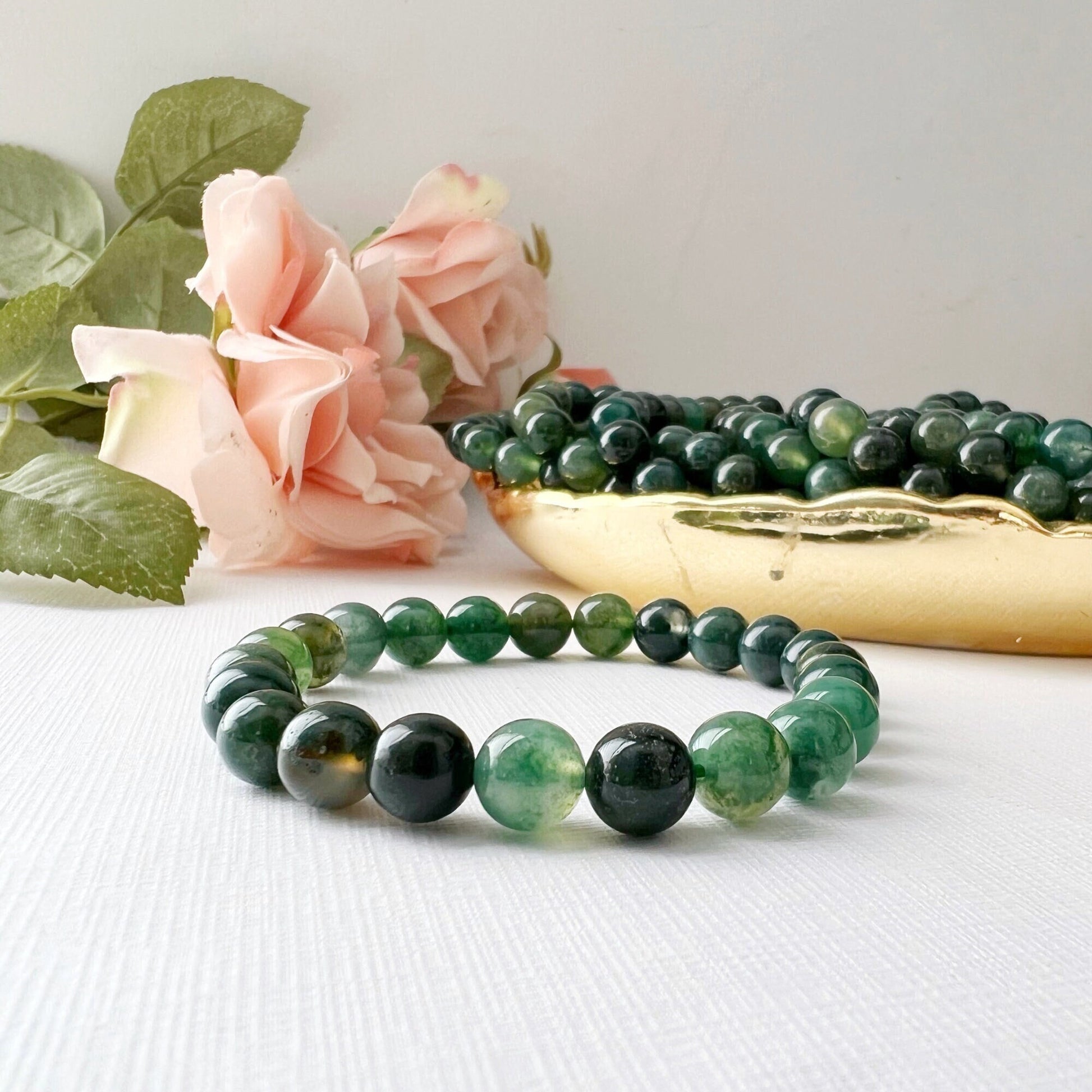 A handcrafted 8mm Green Moss Agate Beaded Bracelet is displayed on a white surface, surrounded by pink roses and green leaves. Behind the bracelet, there's a gold container filled with additional green beads. The well-lit scene highlights the bracelet's metaphysical properties in a minimalist style.