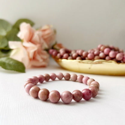 An 8mm Rhodonite Beaded Bracelet is displayed in the foreground on a white surface. Behind it, there is a decorative dish filled with similar pink beads, and a soft-focus bouquet of light pink roses with green leaves in the background, highlighting the metaphysical properties of Rhodonite gemstones.