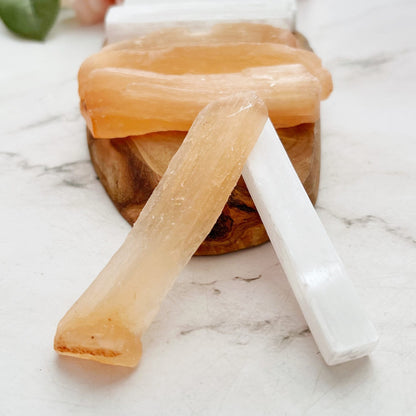 A hand holds two crystal sticks: one is a translucent orange 3" Raw Selenite Stick with rough edges, and the other is a smooth white 3" Raw Selenite Stick. More crystals and green leaves can be seen blurred in the background.