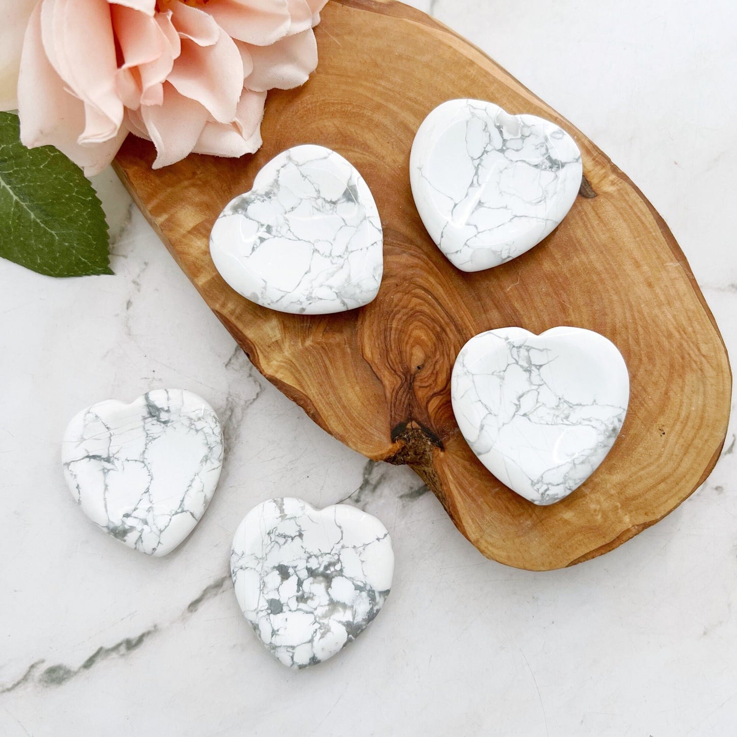 Five white Howlite Heart Shaped Worry Stones with gray veins are placed on a wooden dish on a marble surface, offering calming properties. A light pink rose and green leaf are positioned nearby, adding a touch of color to the serene composition and enhancing stress relief.