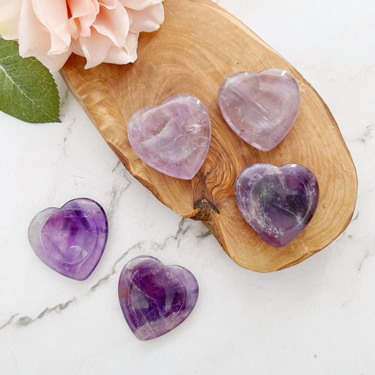 A wooden tray topped with a pink flower, green leaf, and five polished Amethyst Heart Shaped Worry Stones in varying shades of purple rests on a white marble surface, promoting stress relief and emotional balance.