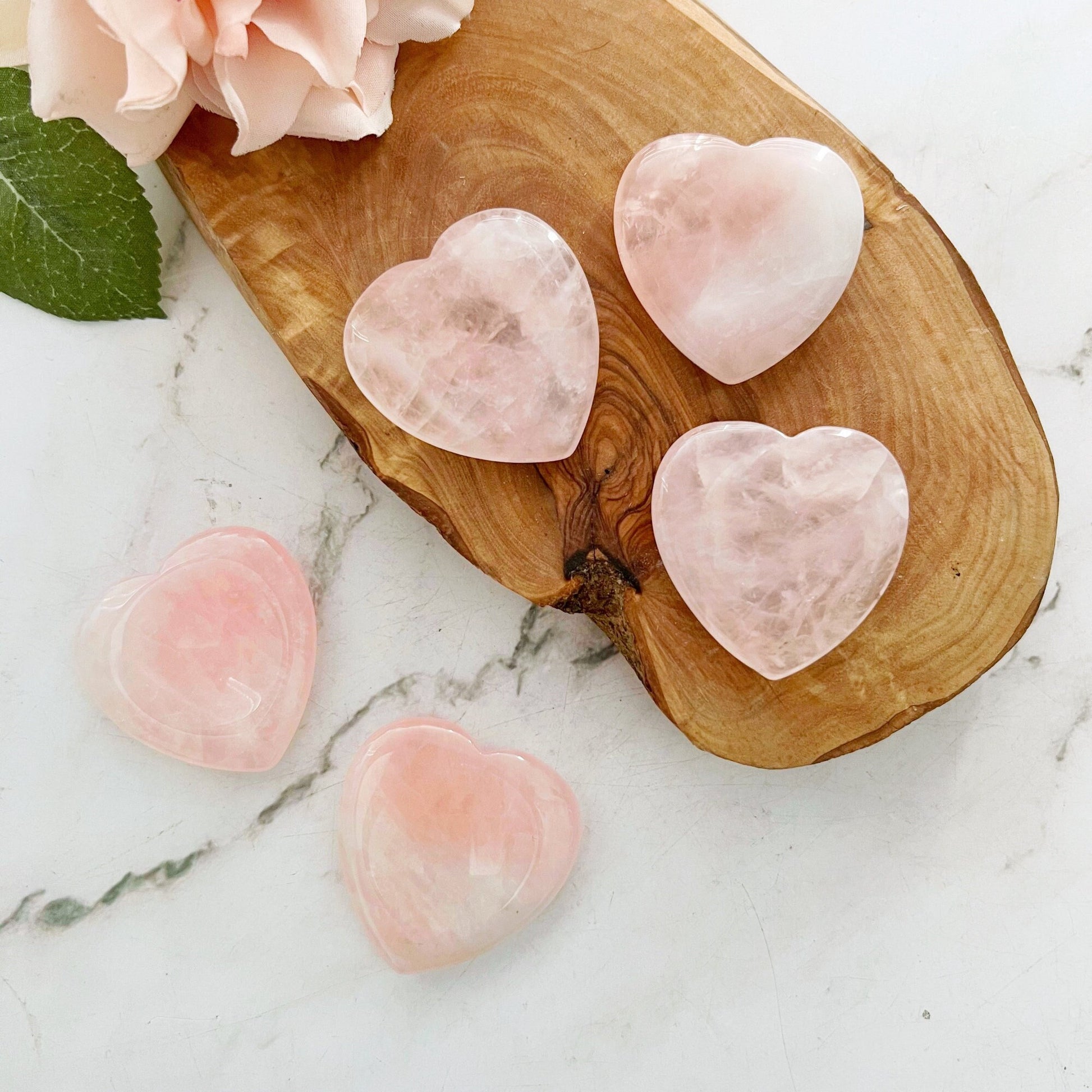 This image shows five pink Rose Quartz Heart Shaped Worry Stones, known for their emotional healing properties, arranged on and around a wooden serving board placed on a marble surface. A peach-colored rose with green leaves is positioned at the top left corner of the wooden board.