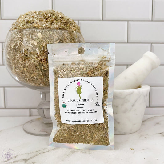 A small, clear bag labeled "Blessed Thistle" from The Stone Sanctuary is displayed in front of a marble mortar and pestle. The bag contains Blessed Thistle, celebrated for its metaphysical properties, while a larger glass container filled with the same herbs sits in the background against a white tile wall.