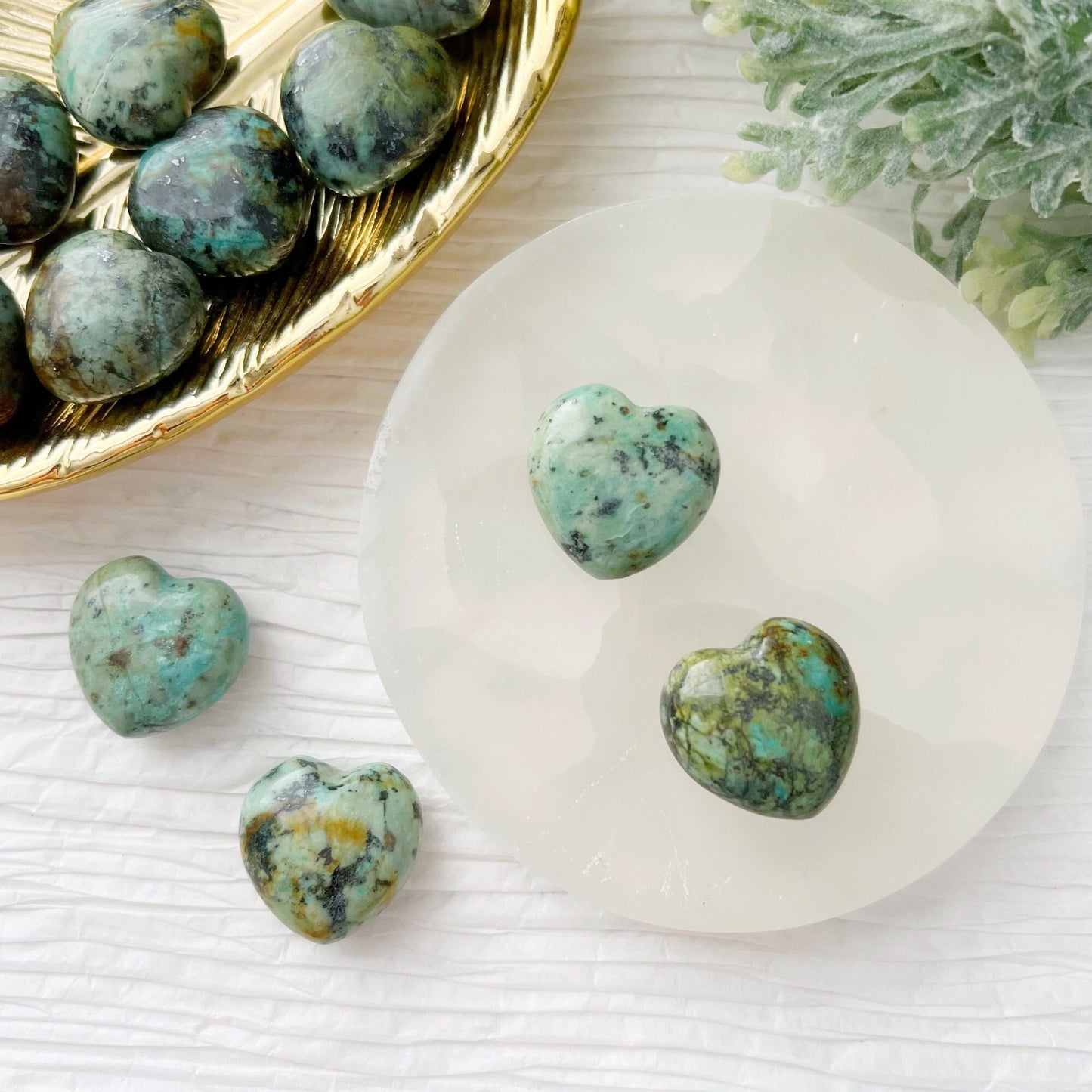 1" Mini African Turquoise Hearts are displayed, with some resting on a white, round, translucent surface and others on a gold dish. Green foliage is partially visible in the background. The heart-shaped gemstones feature a mix of green and darker shades, creating a natural pattern.