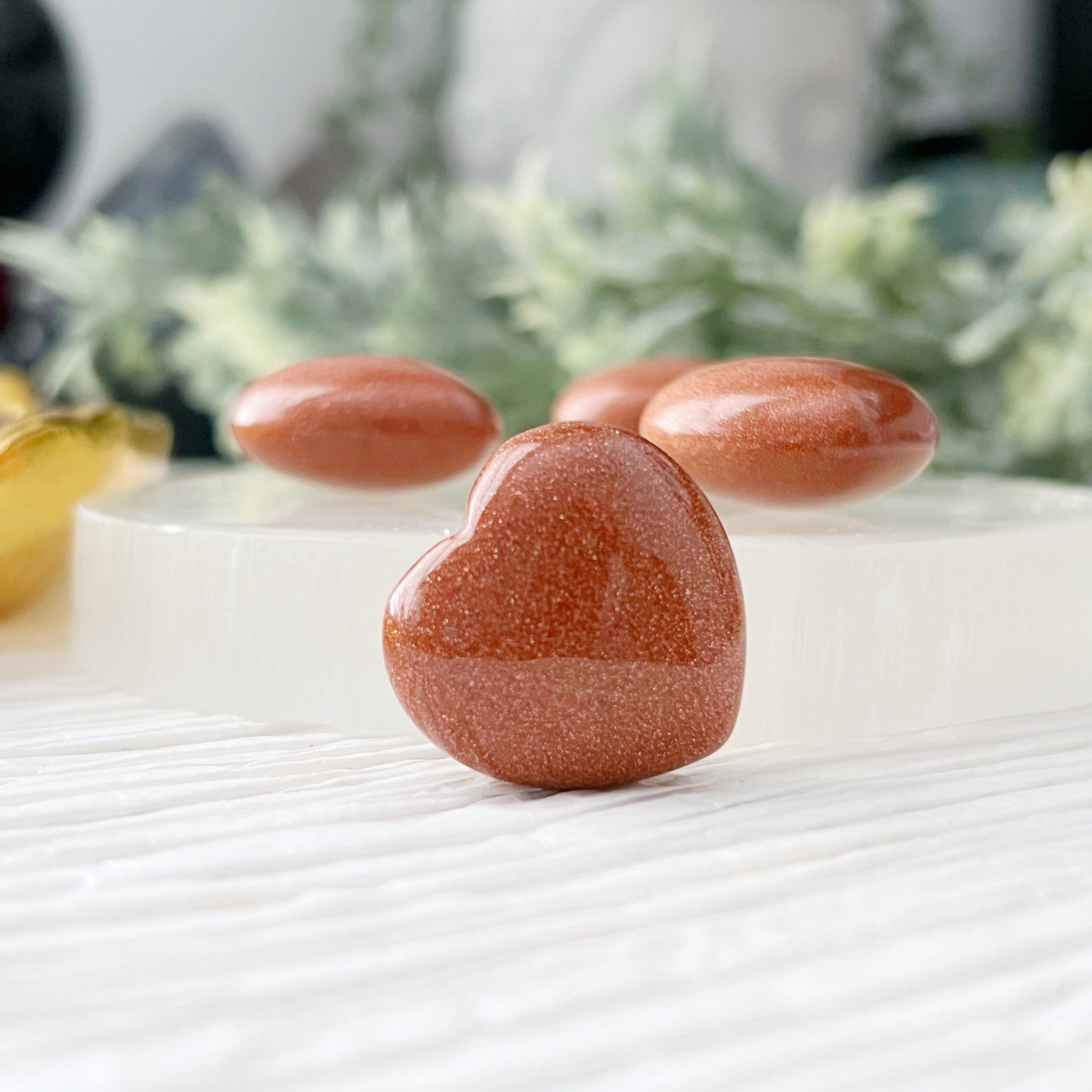 A collection of small, polished heart-shaped gemstones in a reddish-brown color, known as 1" Mini Goldstone Heart, are arranged on a white textured surface. Most rest on a golden dish, with a few displayed on a round white stand. Pocket-sized gemstone accents and green foliage enhance the background.