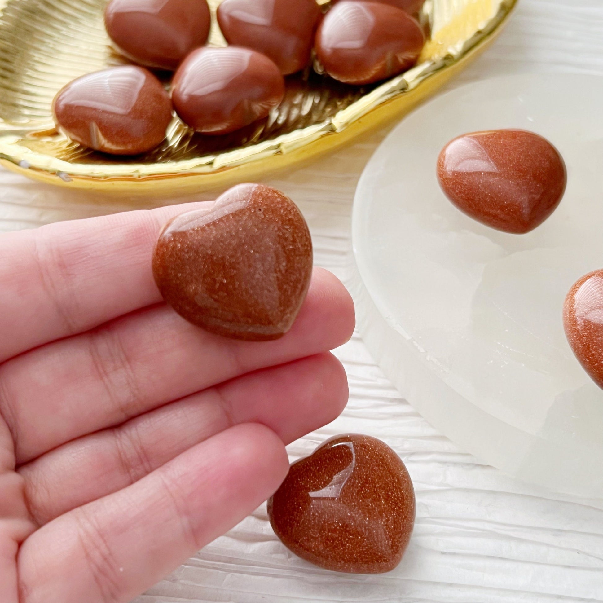 A collection of small, polished heart-shaped gemstones in a reddish-brown color, known as 1" Mini Goldstone Heart, are arranged on a white textured surface. Most rest on a golden dish, with a few displayed on a round white stand. Pocket-sized gemstone accents and green foliage enhance the background.