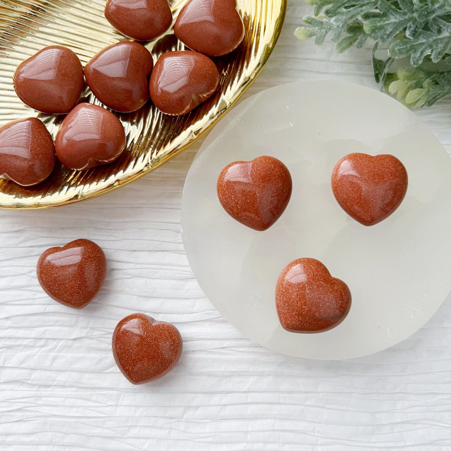 A collection of small, polished heart-shaped gemstones in a reddish-brown color, known as 1" Mini Goldstone Heart, are arranged on a white textured surface. Most rest on a golden dish, with a few displayed on a round white stand. Pocket-sized gemstone accents and green foliage enhance the background.