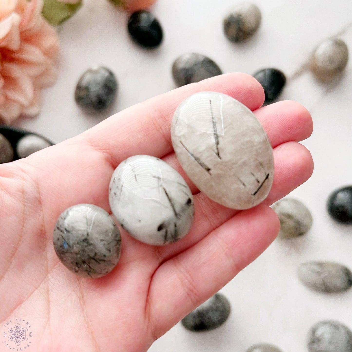 A wooden bowl filled with smooth, polished stones of various shades of grey, black, and white. The stones, including exquisite Black Rutilated Quartz Tumbled Stones, have unique patterns and striations, adding an artistic touch. Known for their metaphysical properties, they contrast beautifully with the bowl’s grain.