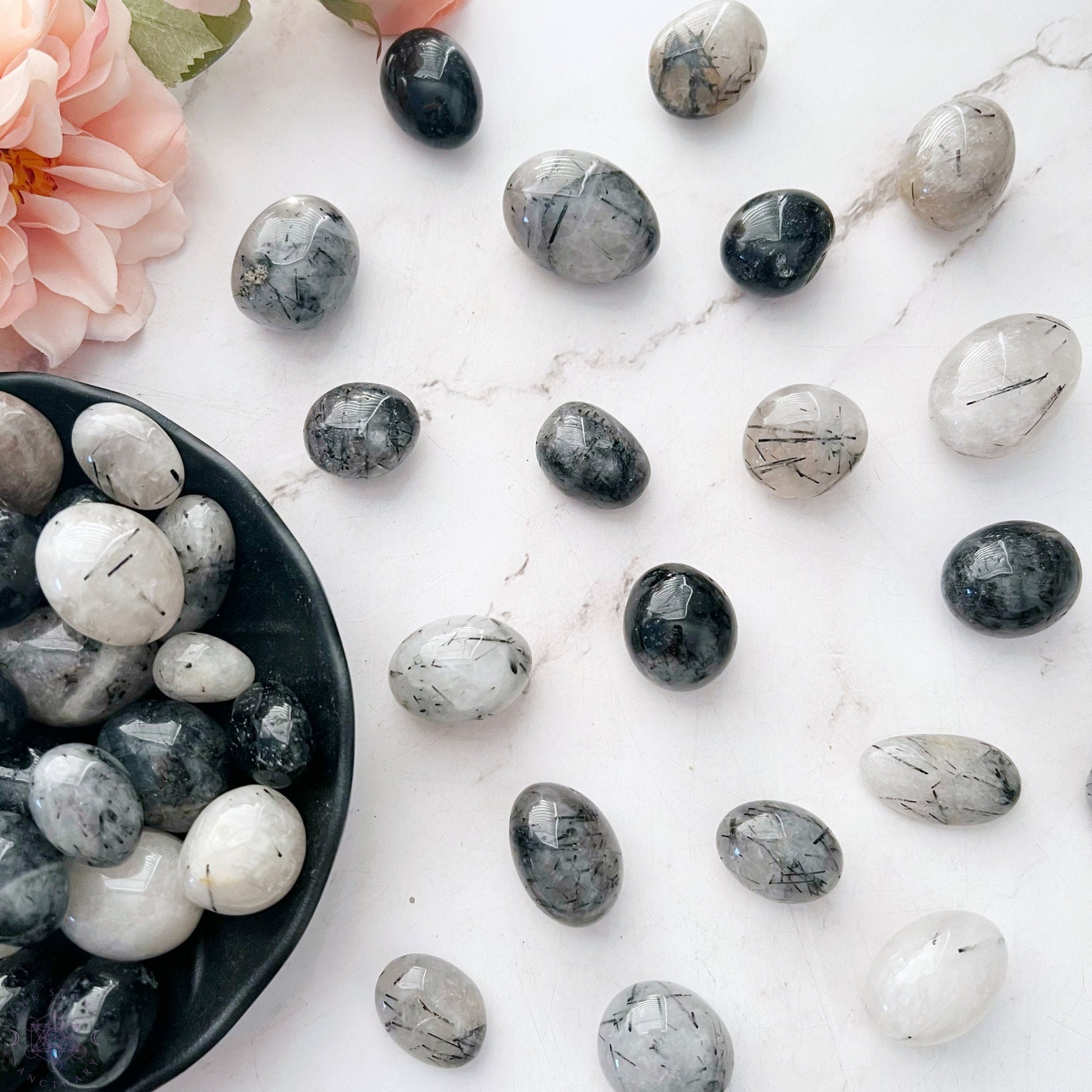 A wooden bowl filled with smooth, polished stones of various shades of grey, black, and white. The stones, including exquisite Black Rutilated Quartz Tumbled Stones, have unique patterns and striations, adding an artistic touch. Known for their metaphysical properties, they contrast beautifully with the bowl’s grain.