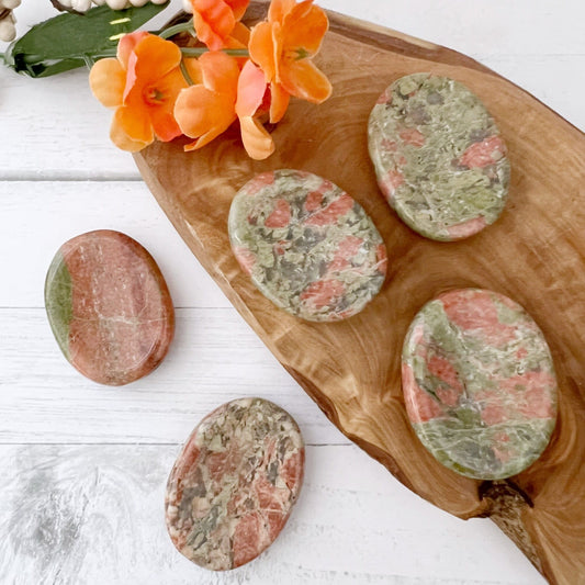 Smooth, oval-shaped Unakite Worry Stones with pink and green marbled patterns are displayed on a wooden slab. To the left, an orange flower with green leaves adds a touch of color to the white wooden background. One Unakite Worry Stone is placed separately on the left side, perfect for emotional healing.