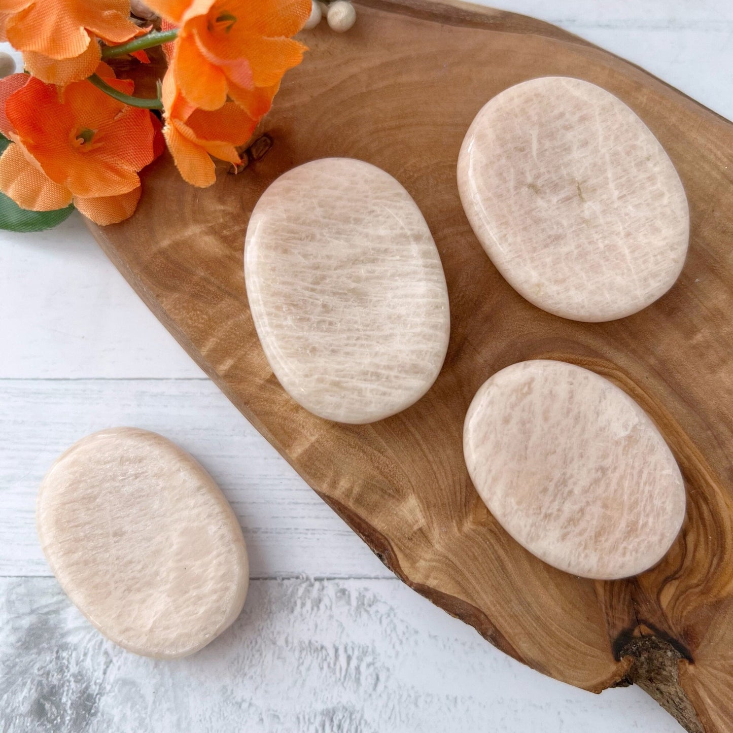 A wooden tray holds four smooth, oval-shaped Peach Moonstone Worry Stones emanating self-loving vibrations. Vibrant orange flowers rest at the top-left corner of the tray. One Worry Stone is placed on the white surface beside the tray, which sits against a background of white plank surfaces.