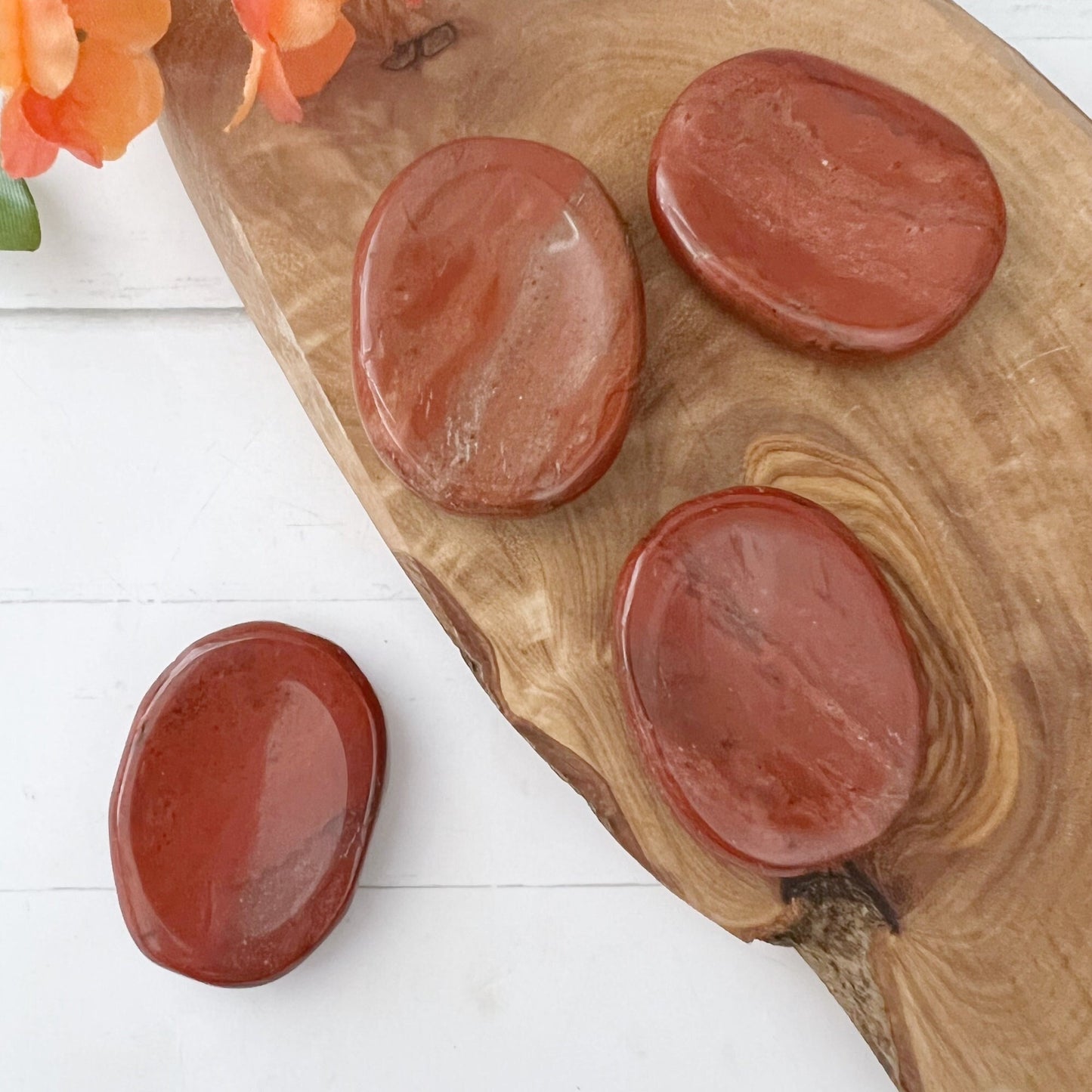 Four smooth, oval-shaped Red Jasper Worry Stones are arranged on a wooden board against a white background. Known for their spiritual energy and connection to the Base Chakra, these stones rest on wood grain patterns with orange flowers in one corner.