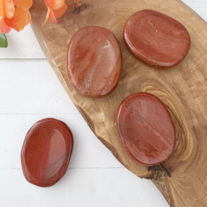 Four smooth, oval-shaped Red Jasper Worry Stones are arranged on a wooden board against a white background. Known for their spiritual energy and connection to the Base Chakra, these stones rest on wood grain patterns with orange flowers in one corner.