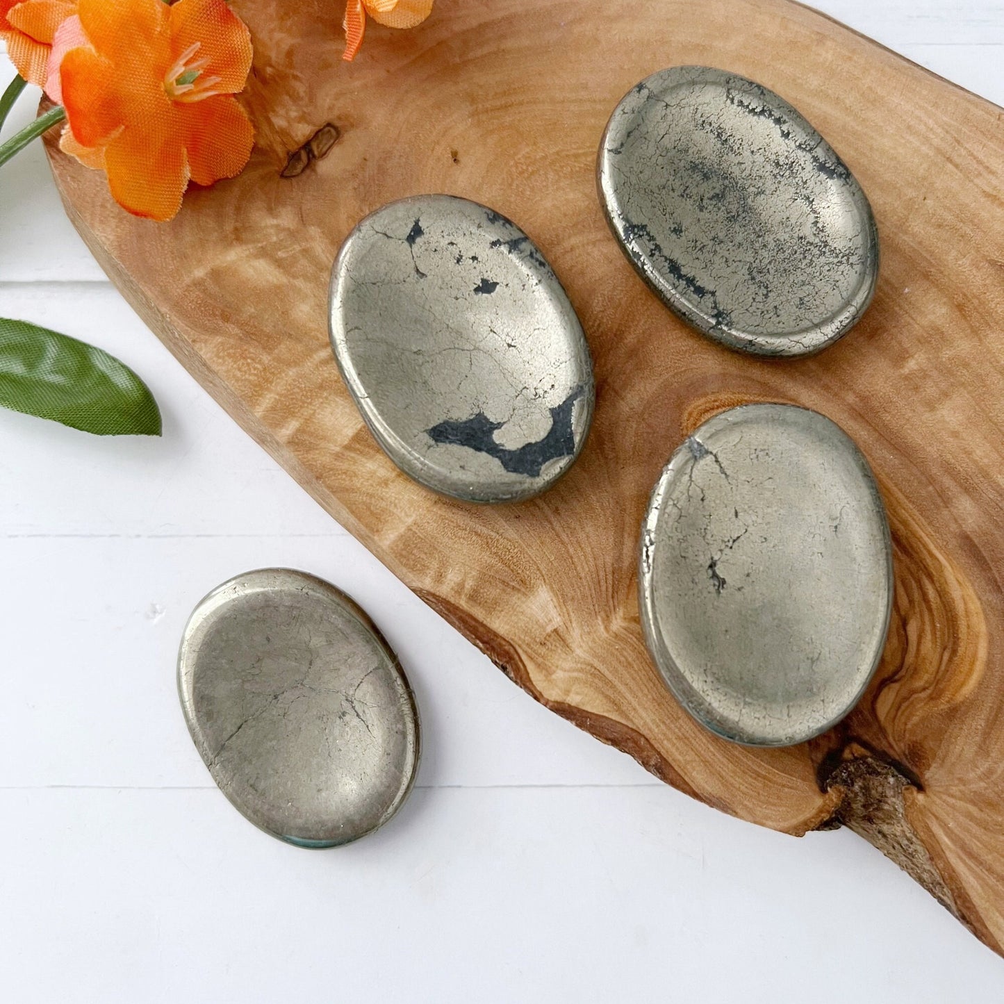 Four polished Pyrite Worry Stones are displayed on a carved wooden board with a couple of vibrant orange flowers placed beside them. The scene is set on a white surface. The stones have a metallic, reflective quality with natural cracks and markings, known for their properties as protection stones.