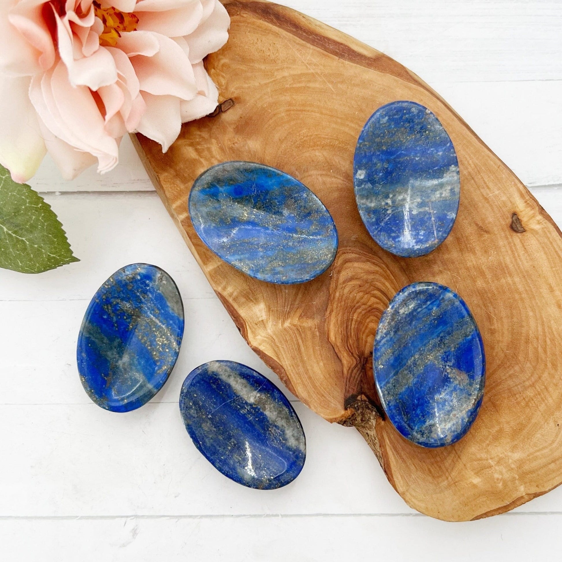 A wooden board holds five polished Lapis Lazuli Worry Stones, known for their spiritual insight and stress relief. A pale pink rose with green leaves is positioned in the top left corner, adding a touch of elegance to the natural stone display. The background is a white wooden surface.