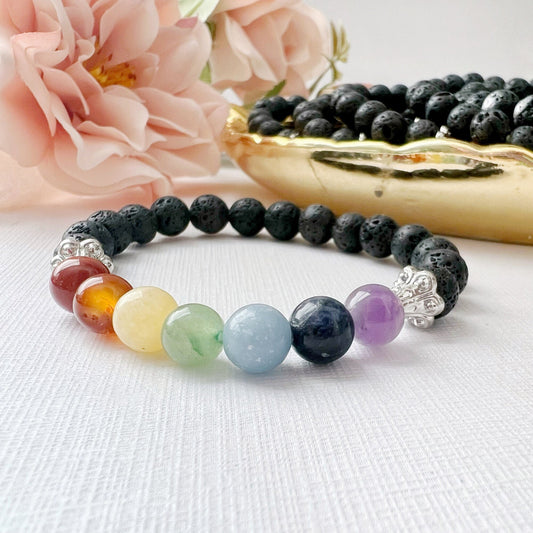 An 8mm Seven Chakra & Lava Rock Beaded Bracelet with black lava stones and seven colored beads, each representing the chakras, is displayed on a white surface. Pink flowers and a golden dish holding additional black beads are in the background, highlighting this chakra healing accessory.