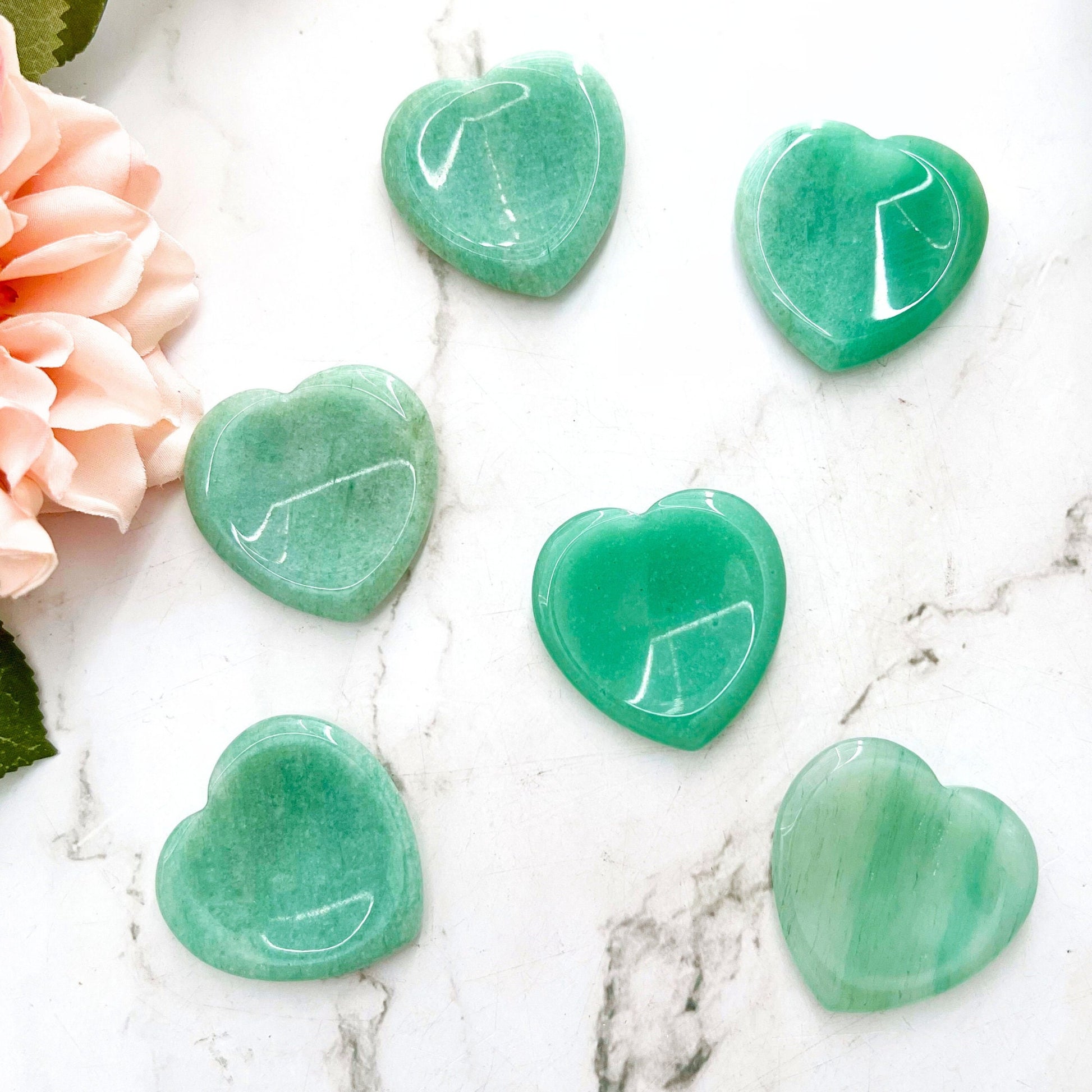 Five polished Green Aventurine Heart Shaped Worry Stones, known for stress relief, are artfully displayed on a wooden board placed on a white marble surface. A pink flower with green leaves is partially visible at the top left corner of the board.