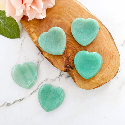 Five polished Green Aventurine Heart Shaped Worry Stones, known for stress relief, are artfully displayed on a wooden board placed on a white marble surface. A pink flower with green leaves is partially visible at the top left corner of the board.