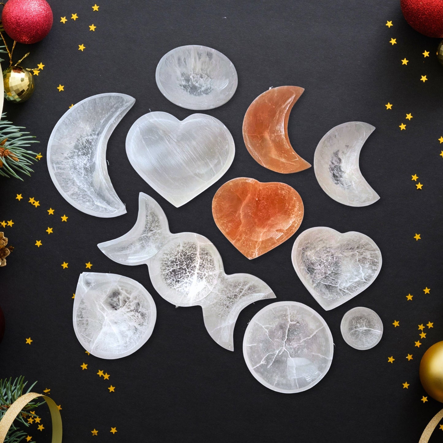 A collection of translucent and opaque stone carvings shaped like moons and hearts, arranged on a black surface scattered with small gold star confetti. Among the shapes are crescent moons and full hearts, primarily white, with one prominent reddish heart. Holiday ornaments, sprigs of greenery, and Selenite Bowl - Select Your Shape and Size surround the scene.