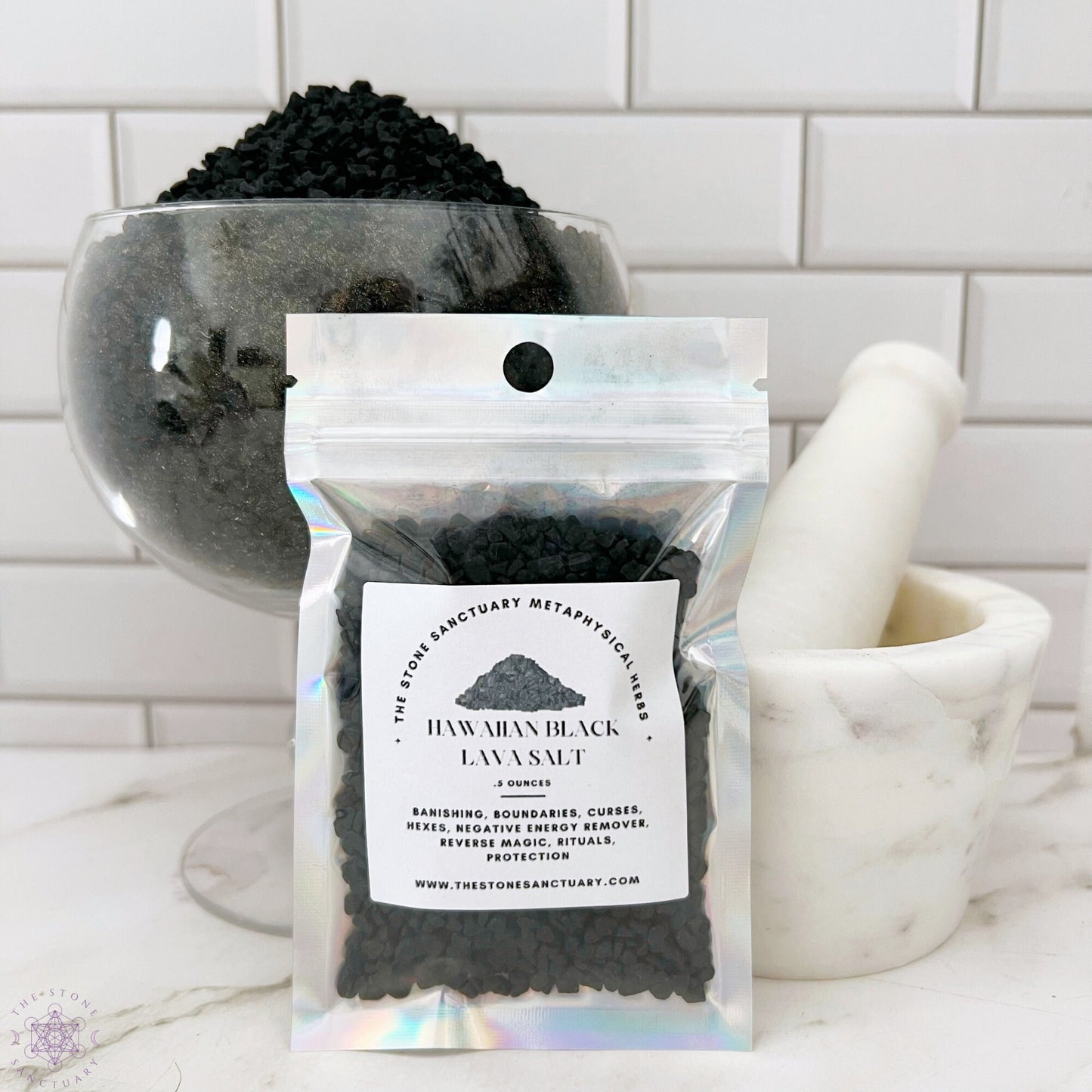 A small sealed package labeled "Black Lava Sea Salt" is placed in front of a glass bowl filled with black salt and a white mortar and pestle. The background features white tiled walls, hinting at the enchanting allure of using Black Salt for Spells with its touch of activated charcoal.