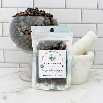 A holographic pouch of Juniper Berries is displayed in front of a clear glass bowl filled with more juniper berries. A white marble mortar and pestle are positioned to the right. The package label mentions metaphysical uses, including anti-theft, curses, and protection to enhance spiritual rituals.