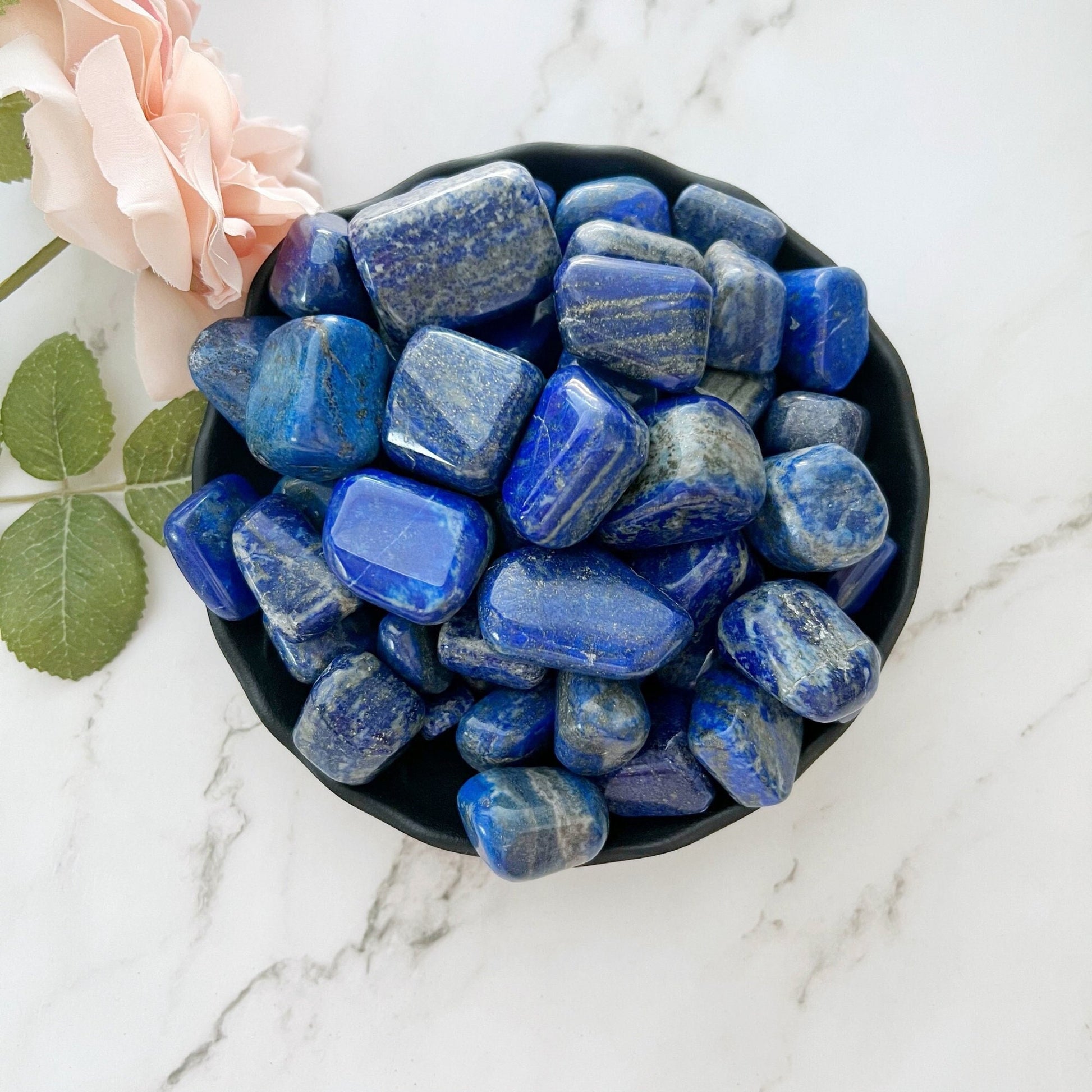 A black bowl filled with polished Lapis Lazuli Tumbled Stones sits on a white marble surface. The stones, valued for their metaphysical properties, are deep blue with occasional gold and white streaks. Next to the bowl is a pink rose with green leaves, symbolizing spiritual growth.