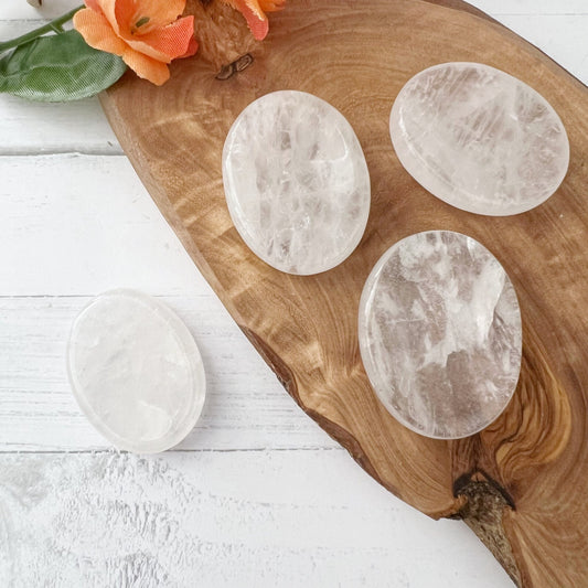 Four polished oval clear quartz crystals are arranged on a wooden board placed on a white surface. Some orange flowers with green leaves are positioned in the upper-left corner of the image. One Clear Crystal Quartz Worry Stone amplifies energy as it sits on the surface beside the board.