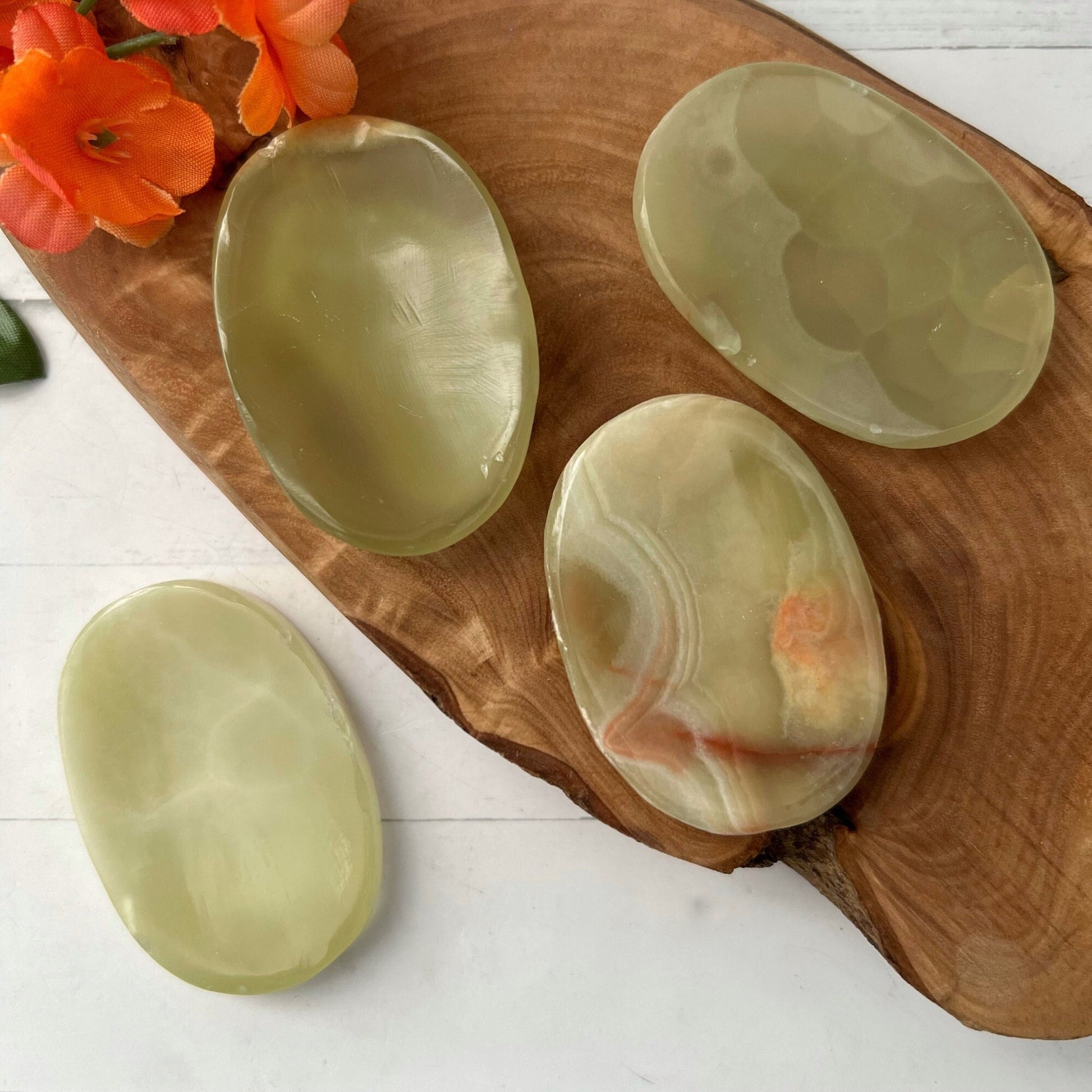 A wooden tray holds four polished, oval-shaped Green Onyx Worry Stones of varying translucency and patterns, known for their heart chakra properties. Orange flowers in the top left corner add a touch of color to the composition. The scene is set on a white surface.