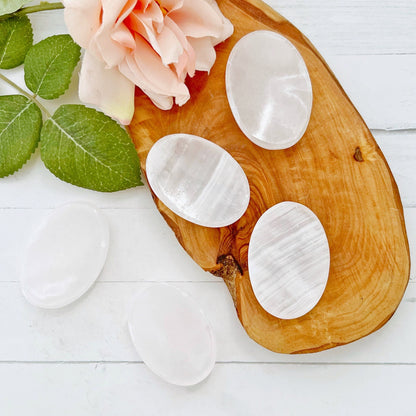 Five smooth, white, oval-shaped stones rest on a wooden tray. Among them lies a Pink Calcite Worry Stone, ideal for stress relief and emotional healing. A light pink rose with green leaves complements the stones. The background features a white wooden surface creating a clean, serene arrangement featuring the Pink Calcite Worry Stone.