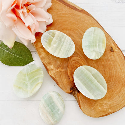 A wooden slab holds five polished **Pistachio Calcite Worry Stones**, arranged neatly. A blooming peach-colored rose with leaves is positioned in the top left corner, adding a touch of floral elegance to the display. The background is a white surface with subtle textures, perfect for stress relief.