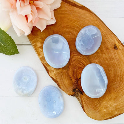 Five smooth, polished Blue Lace Agate Worry Stones are displayed on a wooden slab. A pink flower with green leaves rests in the upper left corner, adding a touch of nature to the composition. Known for stress relief, these worry stones sit elegantly against a clean, white background.