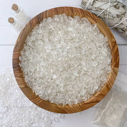 A wooden bowl filled with Clear Quartz Crystal Chips rests on a white surface. Nearby, two small glass bottles with cork stoppers, a bundle of sage tied with string, and a plastic bag partially filled with similar crystal chips complete the serene setting.