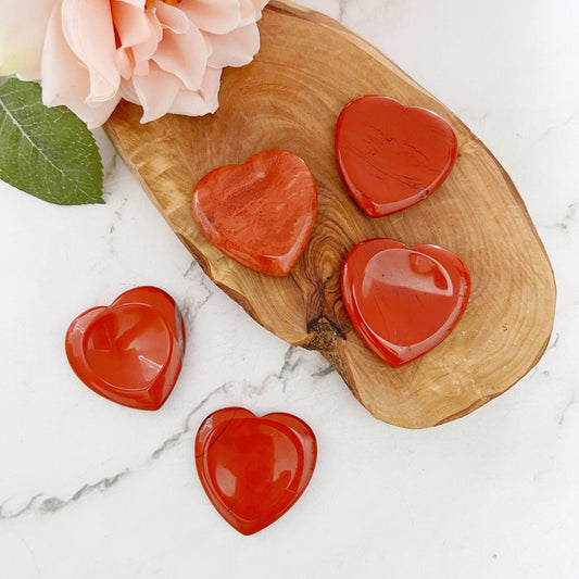 Five polished red heart-shaped stones, including Red Jasper Heart Shaped Worry Stones, are displayed on a wooden tray, surrounded by a white marble surface. The wooden tray rests below a light pink rose with green leaves. Some stones are on the tray, while others are scattered across the marble surface.