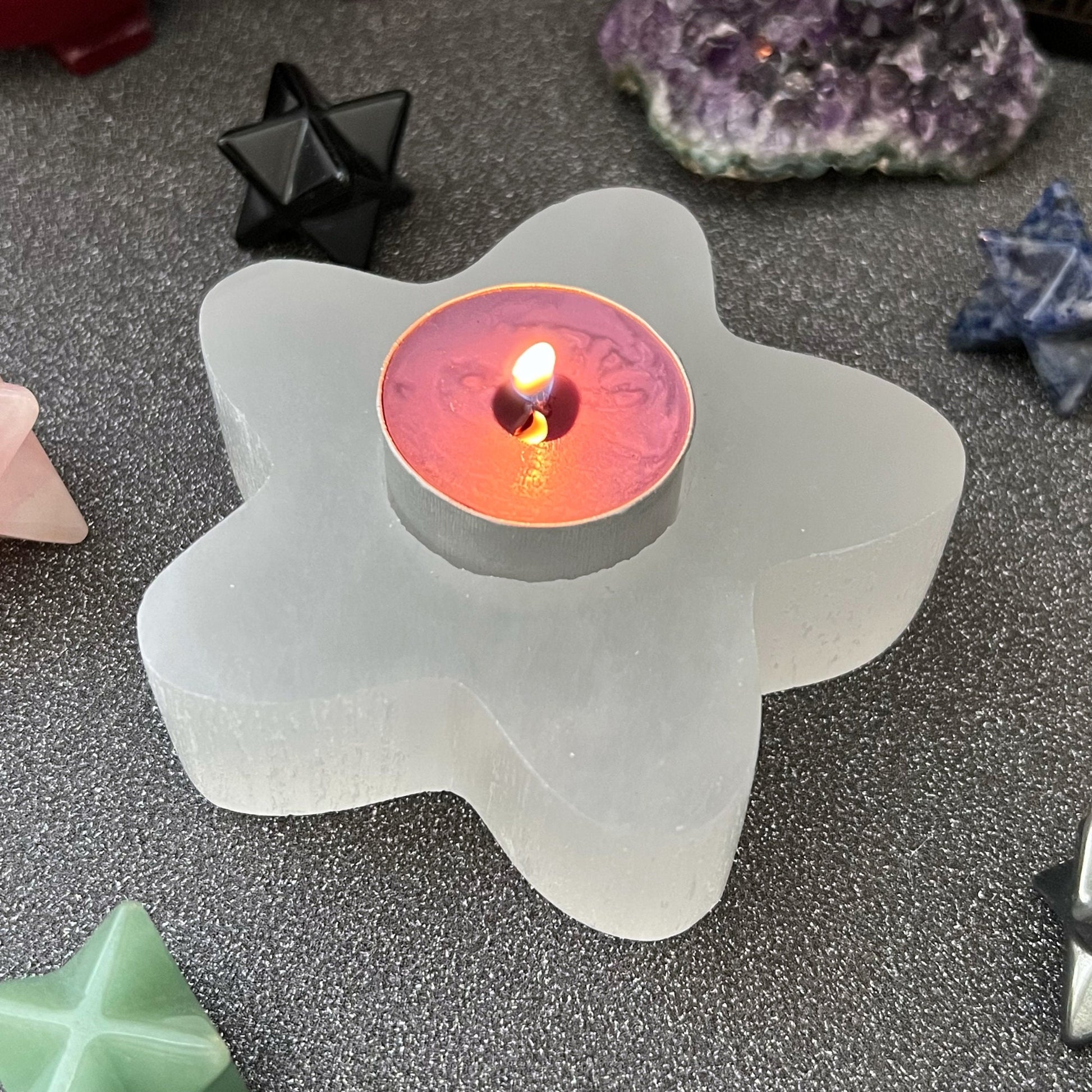 A lit tealight candle sits in a 4" Selenite Star T-Light Holder. The scene is adorned with various crystals and geometric stone figures, including pyramid and star shapes, all displayed on a dark speckled surface—perfect for enhancing your spiritual home decor.