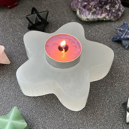 A lit tealight candle sits in a 4" Selenite Star T-Light Holder. The scene is adorned with various crystals and geometric stone figures, including pyramid and star shapes, all displayed on a dark speckled surface—perfect for enhancing your spiritual home decor.