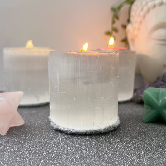 A serene scene features three white candles with soft flames, one prominently in a 2.5" Selenite Cylinder T-Light Holder in the foreground. Surrounding the candles are geometric crystals and a partial view of a Buddha statue in the background. The setting evokes spiritual clarity and meditation.