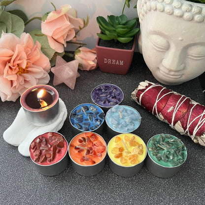 An arrangement of the Set of 7 Unscented Chakra Tealight Candles in small silver tins is displayed on a table with an unscented colored tealight candle burning next to them. Surrounding items include a potted plant, pink flowers, a "Dream" decoration, a Buddha head sculpture, and a sage bundle.